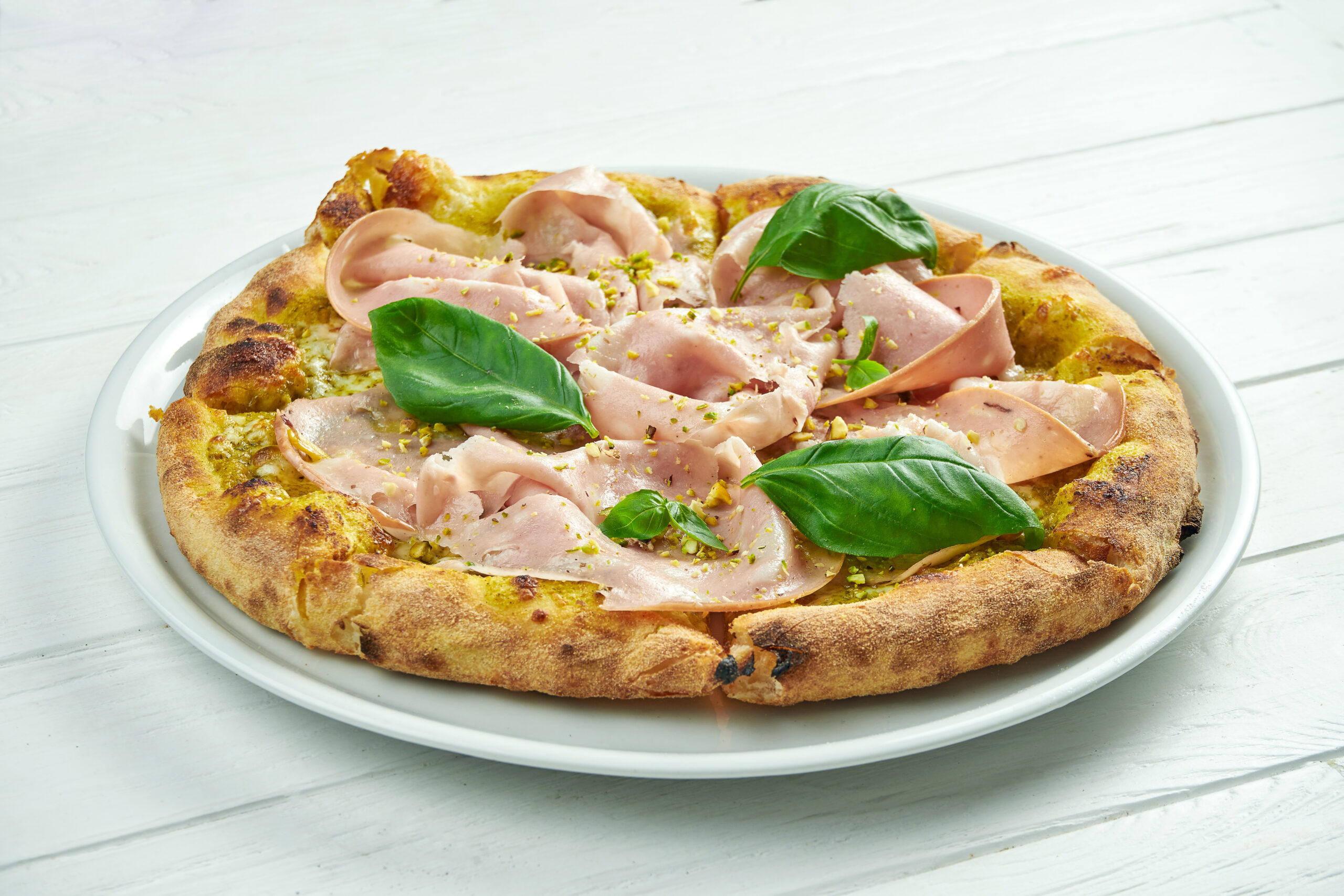 Appetizing homemade pizza with mortadella sausage, pistachios, basil and melted cheese on a white plate on a white wooden background